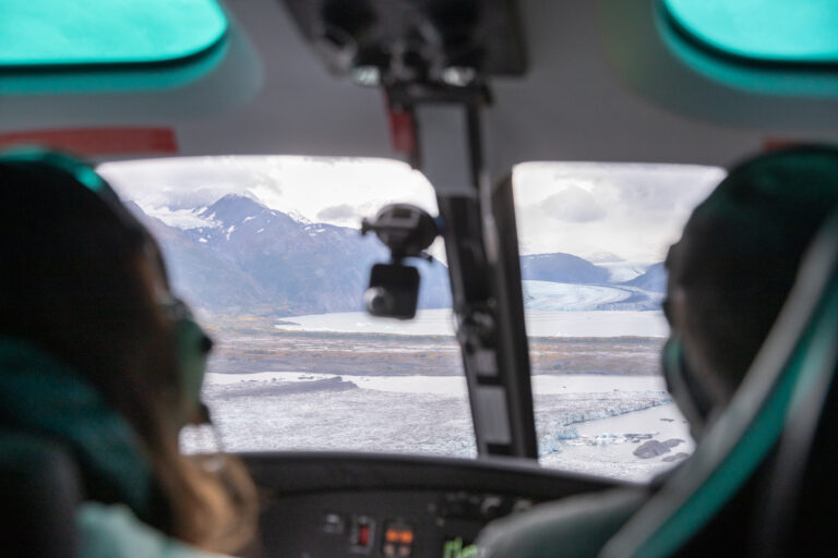 Two people sit in the front seat of a helicopter as they look out the window at the mountains and glaciers.