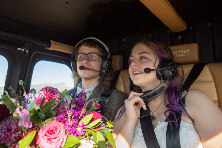 A bride and groom sit inside a helicopter with headphones on and a bouquet of flowers on their laps.