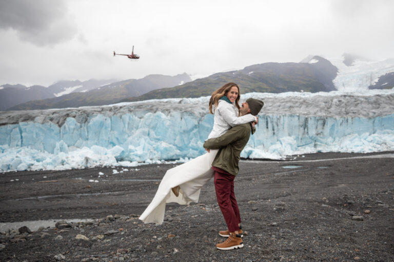 A groom holds his bride up in the air as they stand in front of a blue glacier in Alaska and a helicopter flies in the sky behind them.