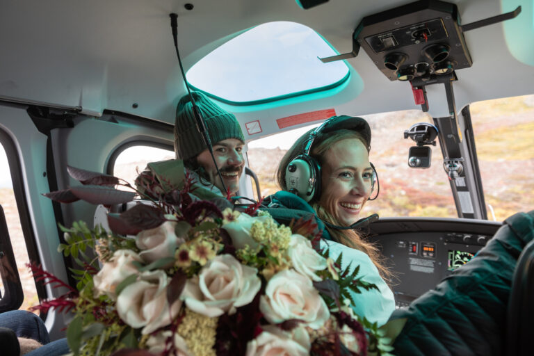 A bride and groom sit in the front seat of a helicopter smiling back at the camera