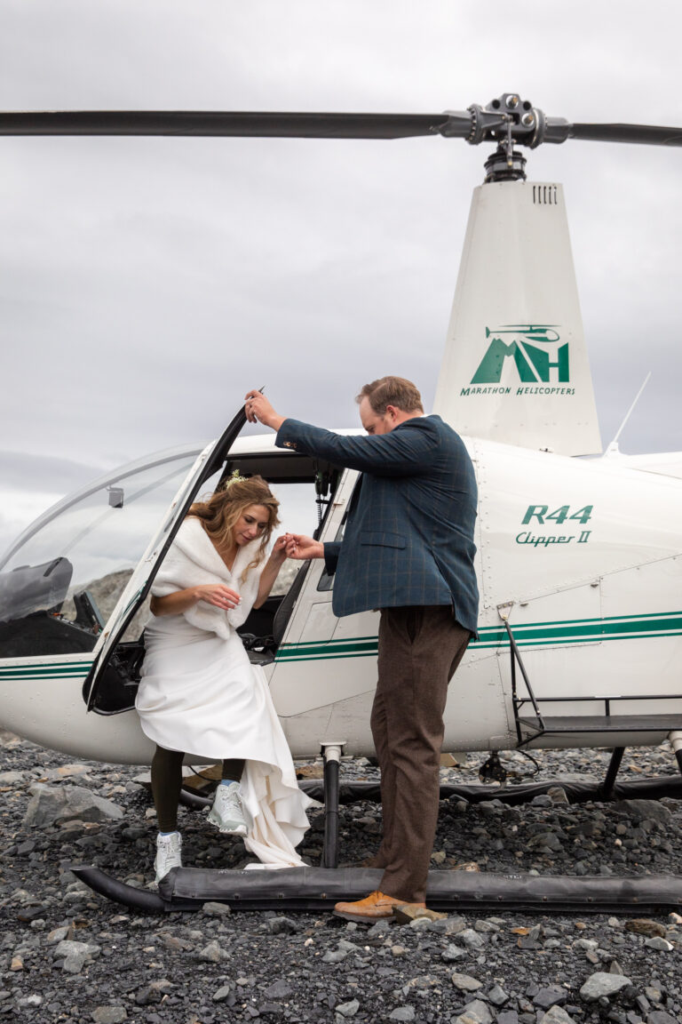 A groom helps his bride out of a helicopter on their elopement day in Alaska.