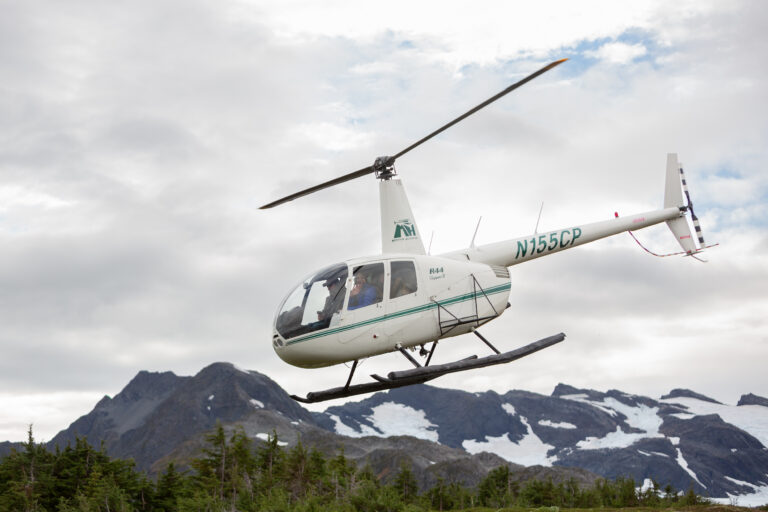 A woman in a blue jacket waves out of a helicopter window as it flies into the sky in Alaska.