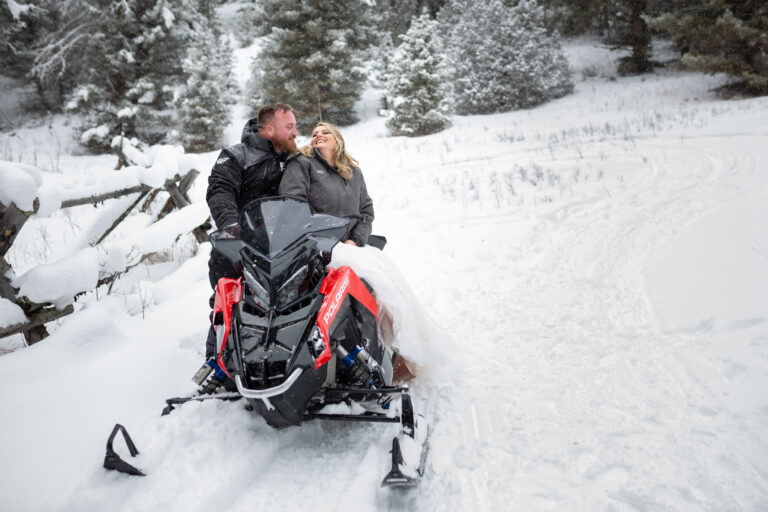 A bride sits on a snowmobile while her groom stands behind her on their elopement day in Montana.