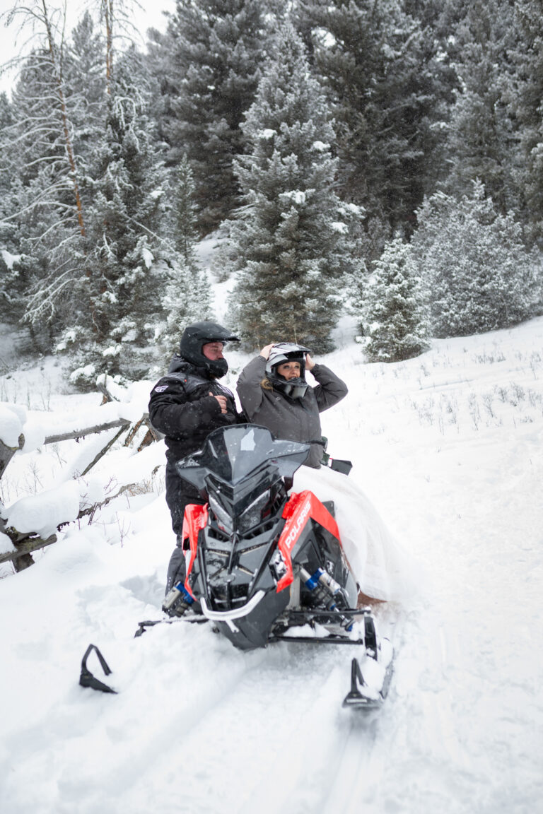 A bride sits on a snowmobile putting her helmet on as her groom stands behind her adjusting his snow suit on their winter elopement in Montana.