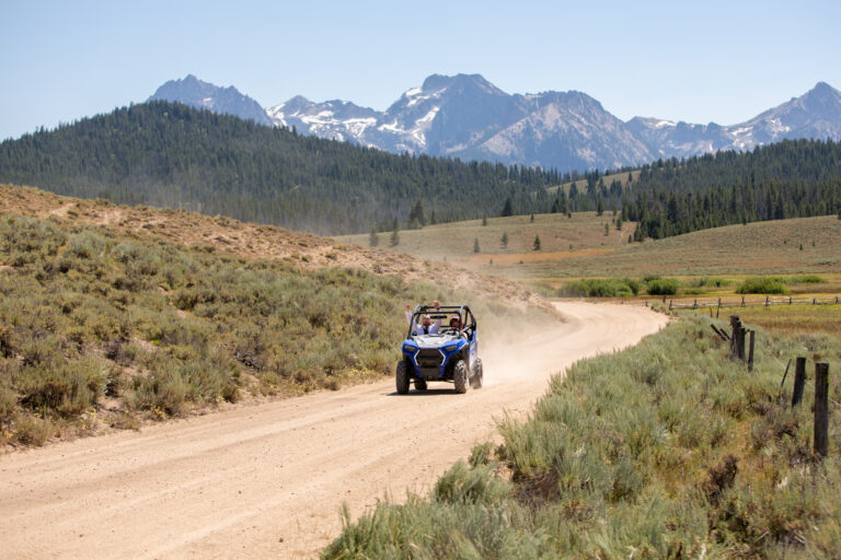A bride and groom ride in a side-by-side on a dirt road with mountains behind them in Stanley Idaho.
