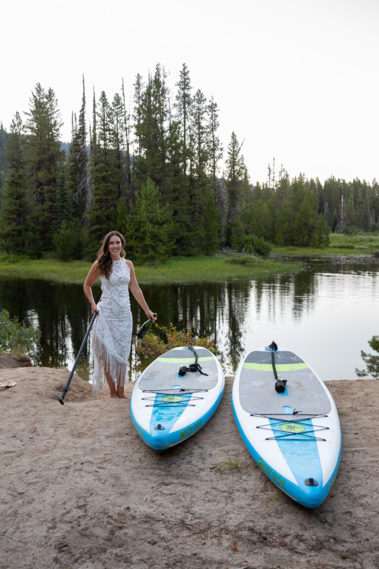 A bride walks up a sandy beach towards to paddle boards, holding a bottle of champagne in one hand and a paddle in the other