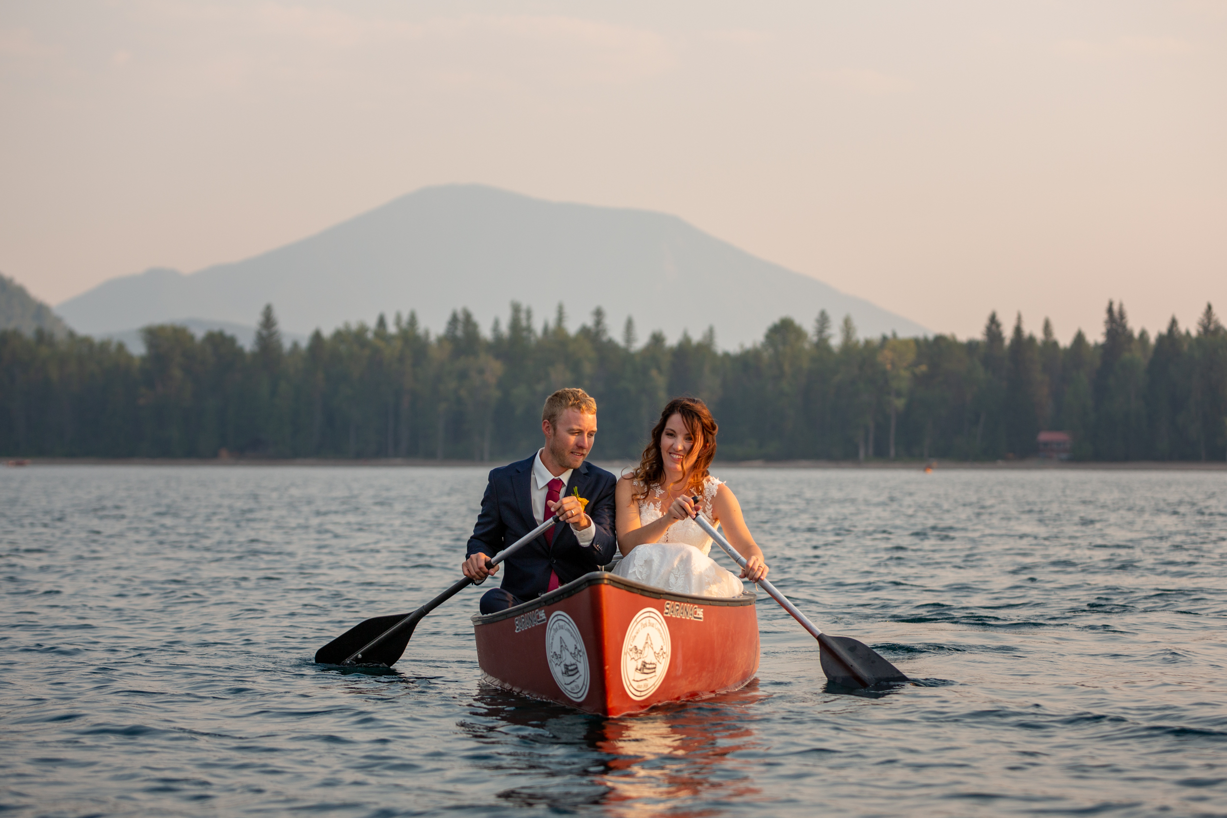 A bride and groom sit side by side in a canoe paddling across Lake McDonald in Glacier National Park.