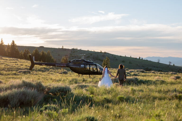 A bride and groom walk hand in hand through a meadow towards a helicopter.