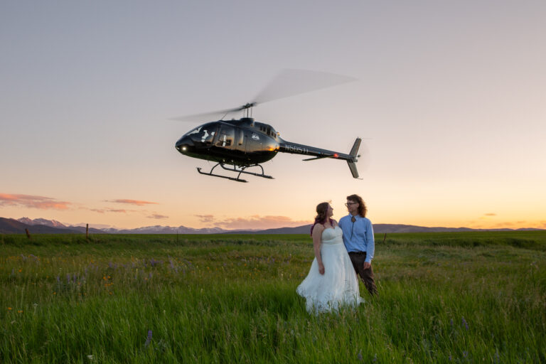 A bride and groom stand smiling at each other in a grassy field of wildflowers as a helicopter takes off into the sky behind them.