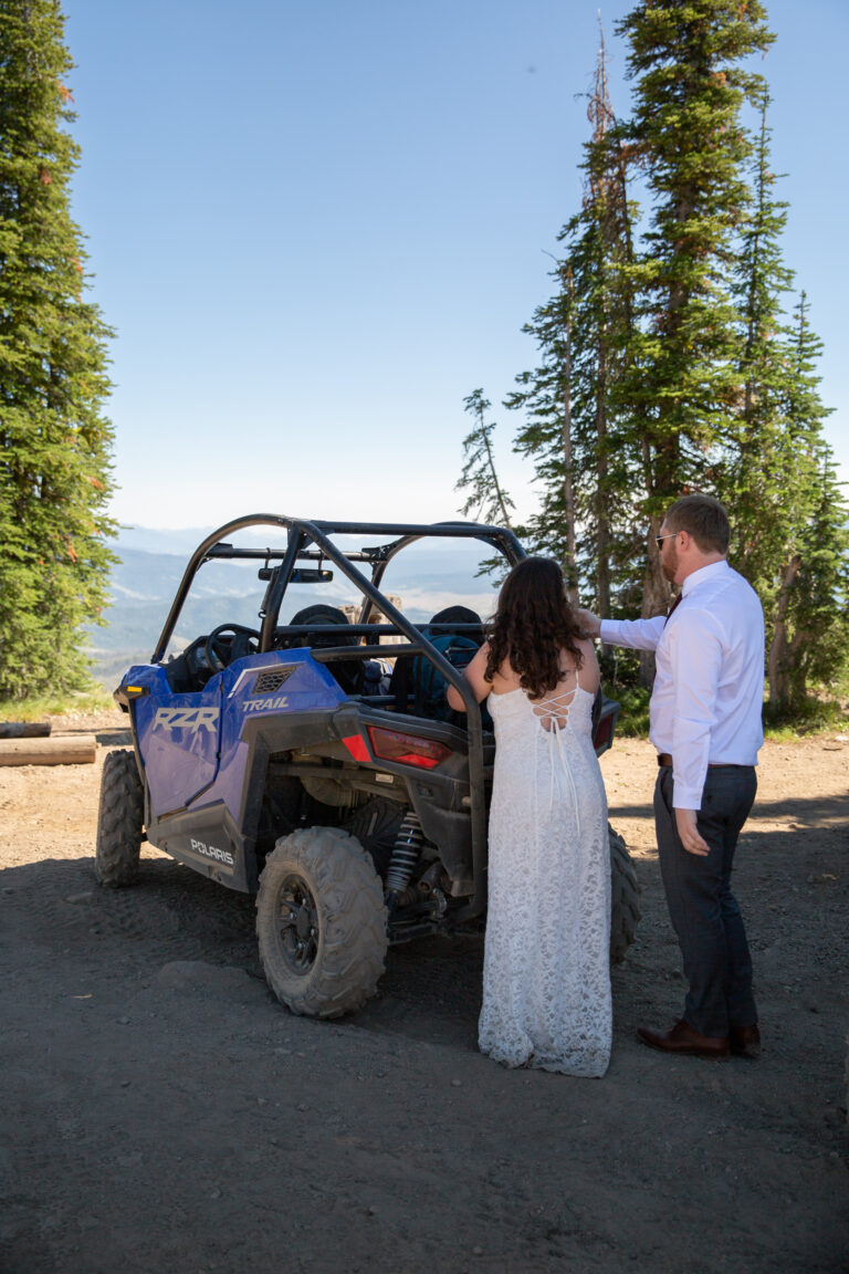 A bride and groom stand behind a parked side-by-side while looking at the mountain views around them in Idaho on their elopement day.