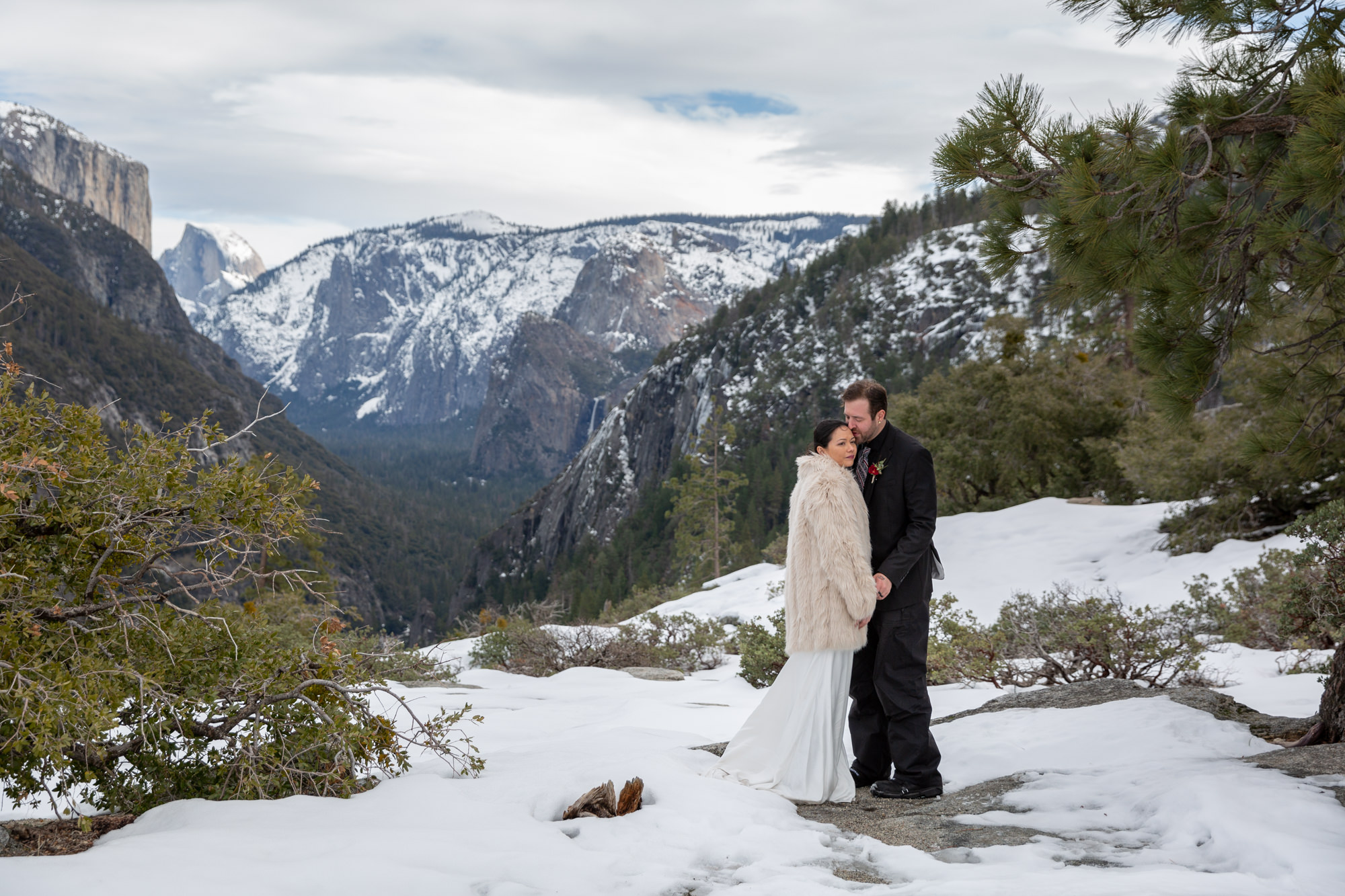 A bride in white dress and white fur coat leans against her groom smiling after they decided to have an elopement versus a wedding in Yosemite.