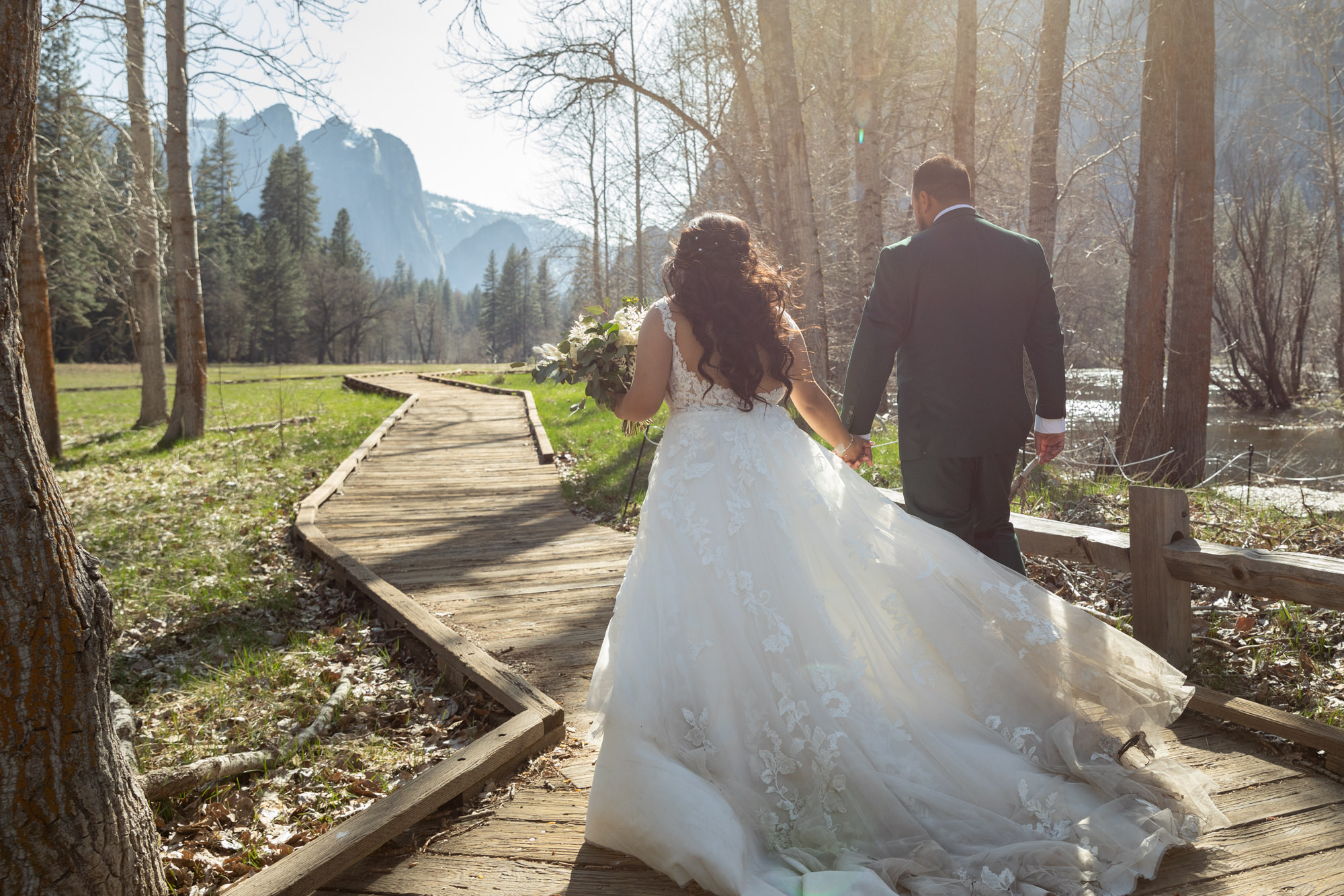 A bride and groom walk hand in hand down a wooden boardwalk in Yosemite as her elopement wedding dress blows in the wind.