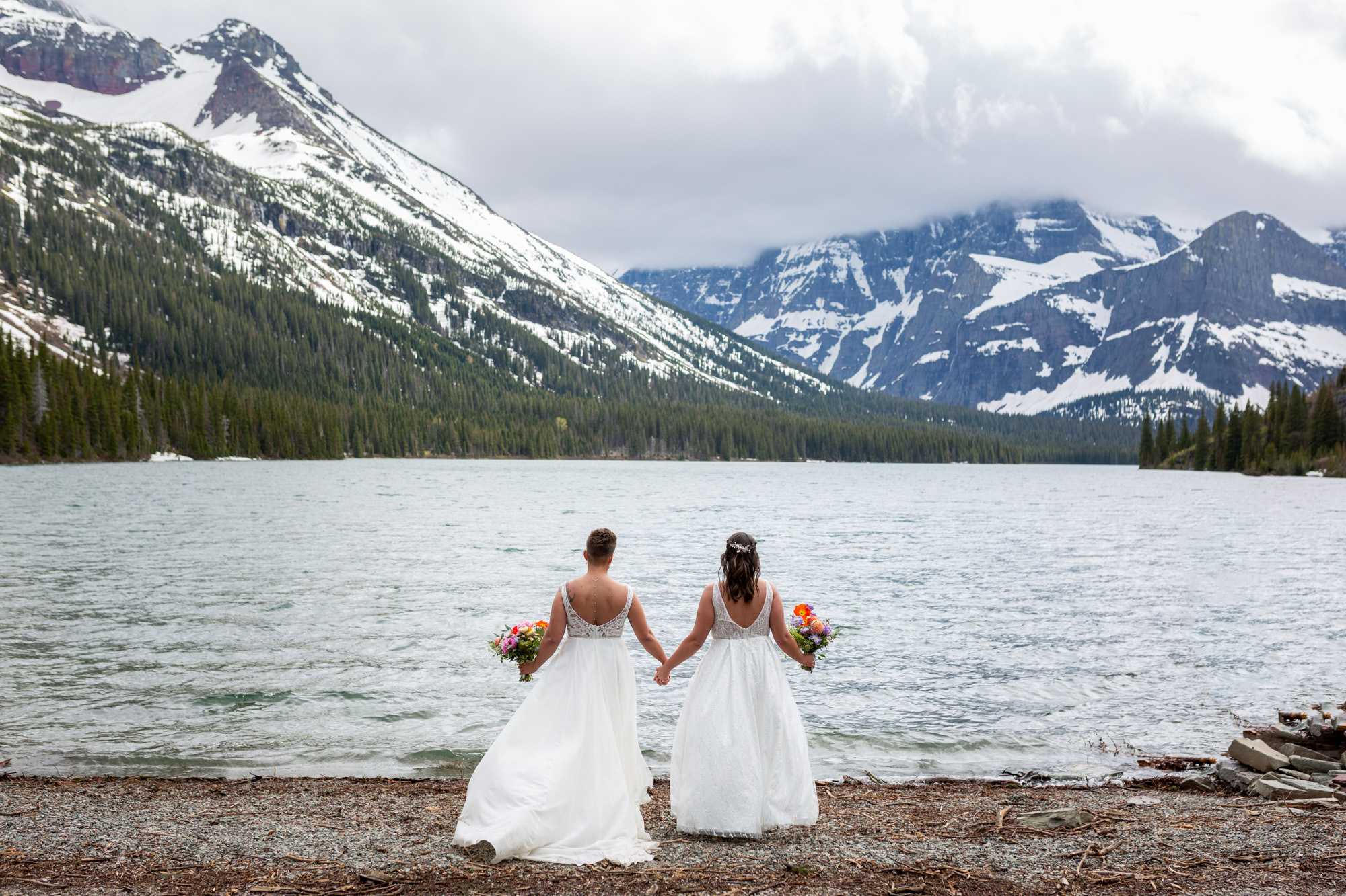 Two brides stand holding hands on the shoreline of Lake Josephine in Glacier National Park.