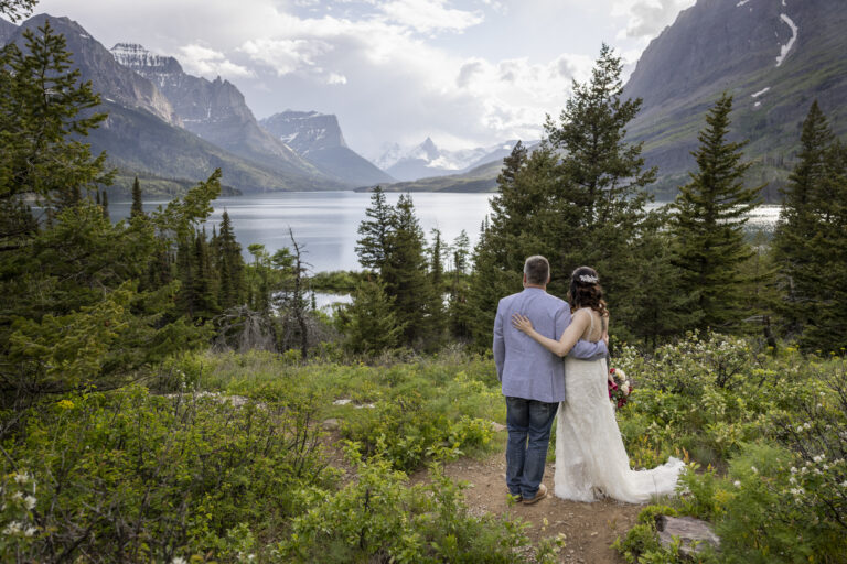 Eloping in Glacier National Park: Your Complete Guide | Scenic Vows A bride and groom stand surrounded by green plants and wildflowers staring out over a lake at the mountains around them in Glacier National Park