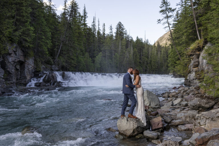 Eloping in Glacier National Park: Your Complete Guide | Scenic Vows A bride and groom stand on a rock next to a waterfall, kissing with tress surrounding them.