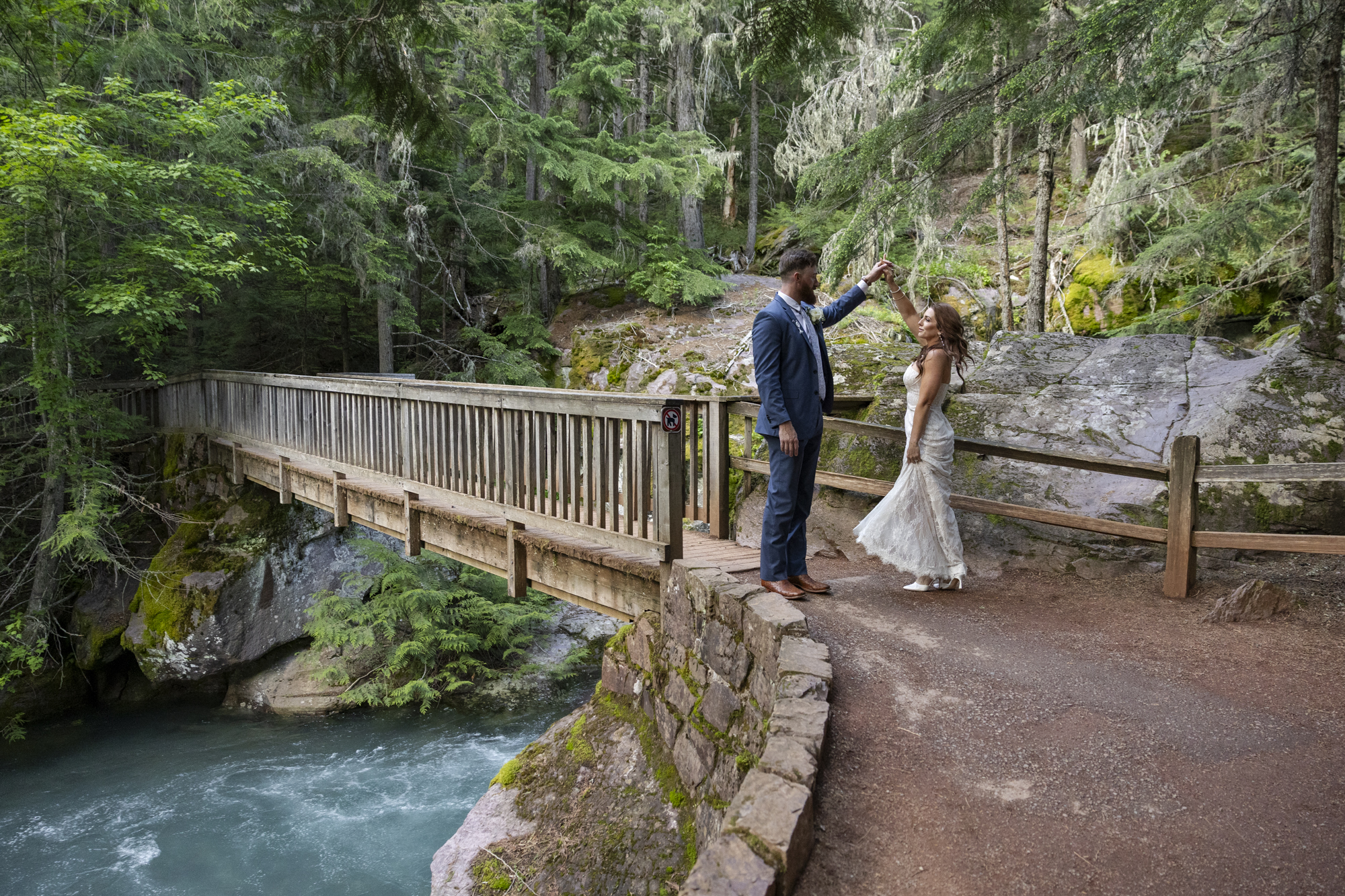 Eloping in Glacier National Park: Your Complete Guide | Scenic Vows A bride and groom stand at the end of a bridge as the groom spins the bride around and they laugh.