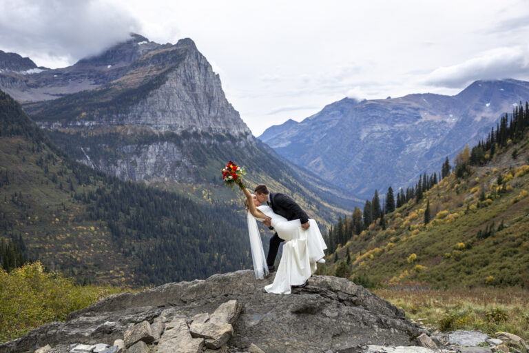Eloping in Glacier National Park: Your Complete Guide | Scenic Vows A bride and groom stand on a rock as the groom dips the bride back as they kiss and she holds her colorful bouquet of flowers in the air
