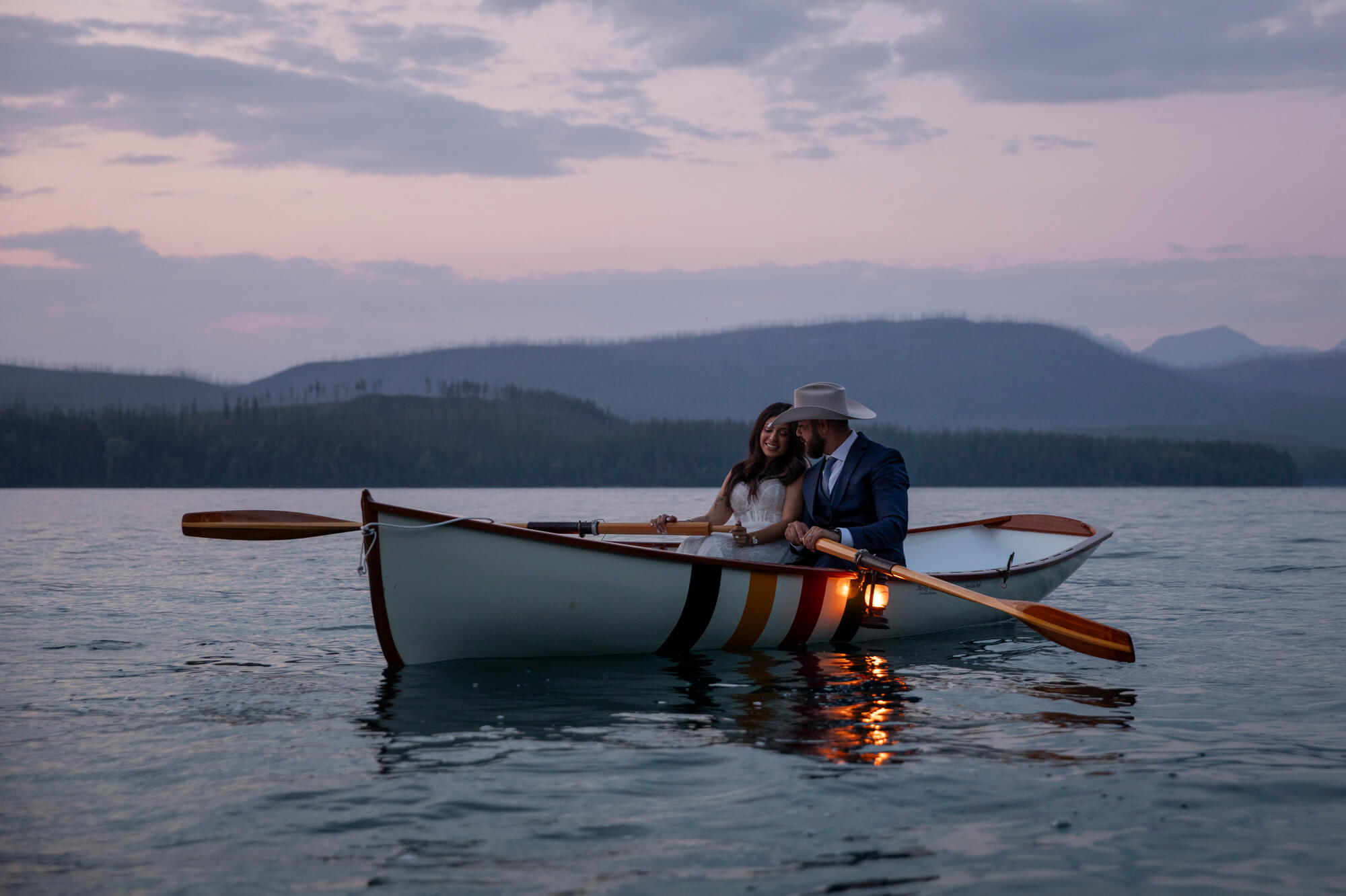 Eloping in Glacier National Park: Your Complete Guide | Scenic Vows A bride and groom sit side by side in a canoe on a lake at sunset with a lantern hanging on the side of the canoe.