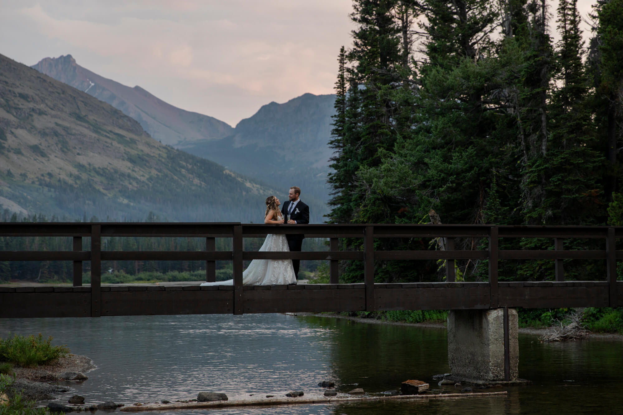 Eloping in Glacier National Park: Your Complete Guide | Scenic Vows A bride and groom stand on a bridge over a lake surrounded by pine trees and mountains in the background.