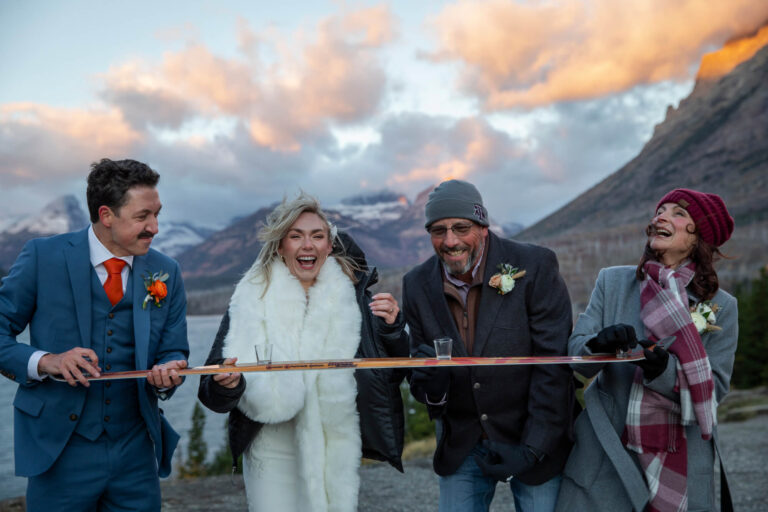 Eloping in Glacier National Park: Your Complete Guide | Scenic Vows A bride and groom along with two guests hold a shotski and laugh as the sunrises behind them.