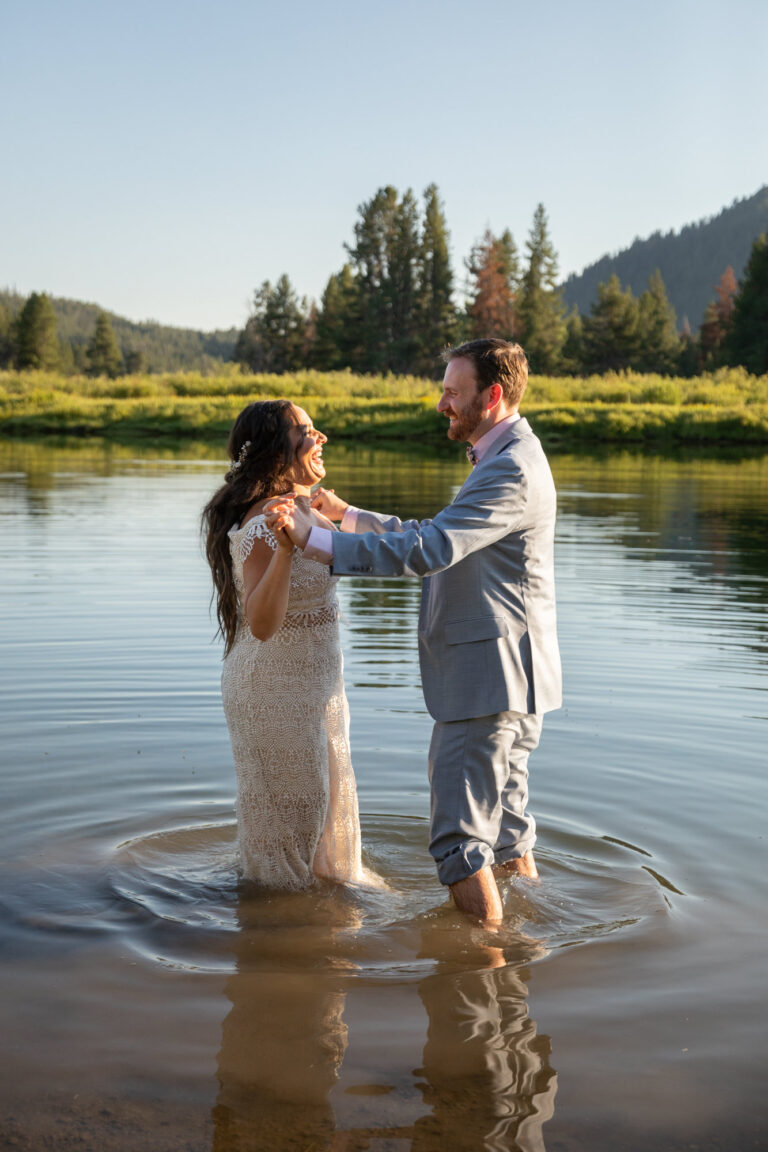 A bride and groom hold hands and laugh while standing in a river after eloping in Grand Teton.