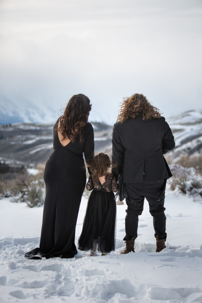 Two brides, wearing black, stand holding hands with a little girl who stands between them.
