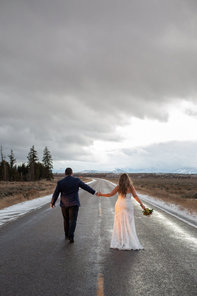 A bride and groom walk down a road holding hands after eloping in Grand Teton.