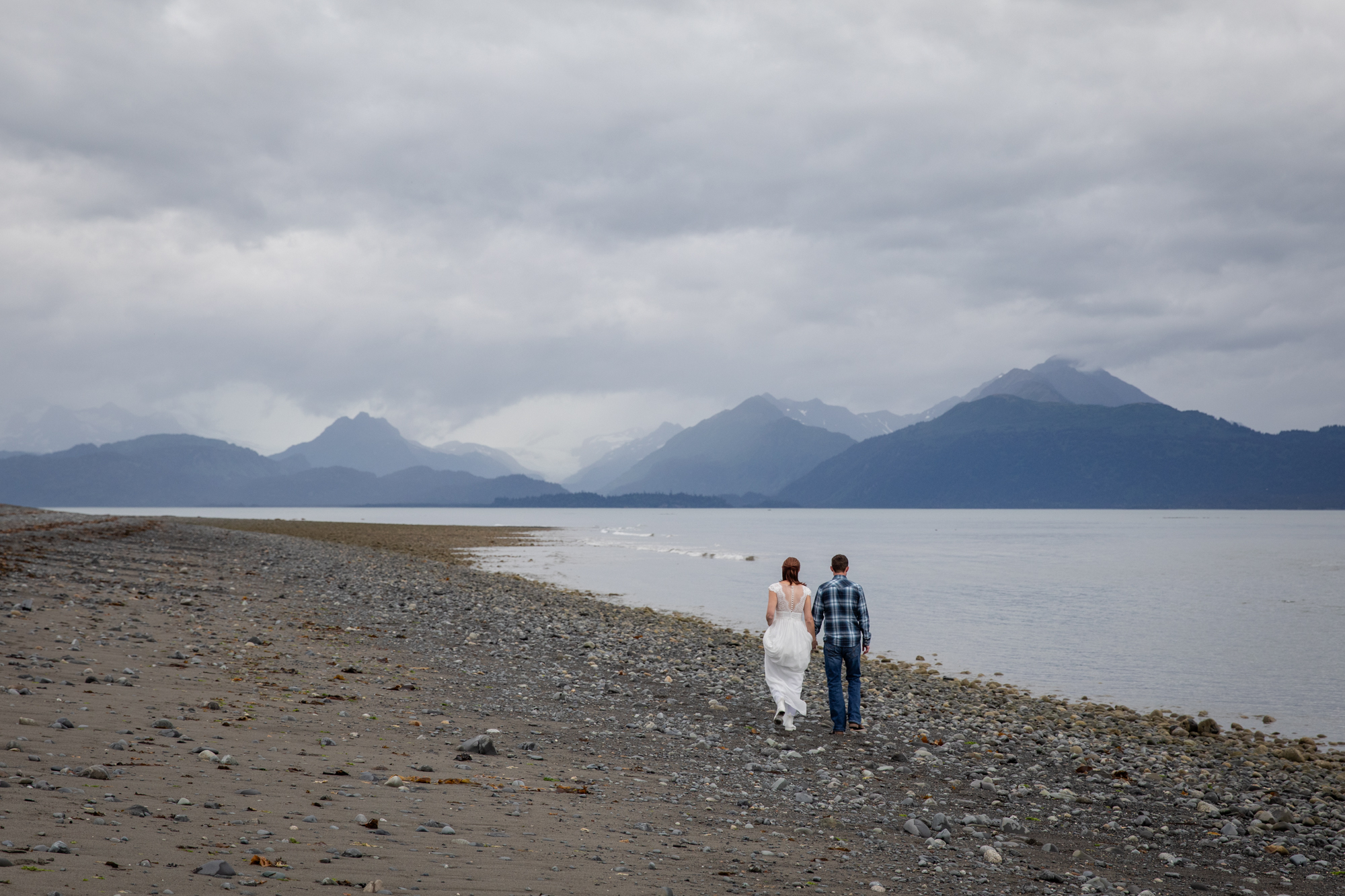 Couple walking along a quiet beach near Kachemak Bay, Alaska with expansive ocean views and distant mountains during an intimate elopement.