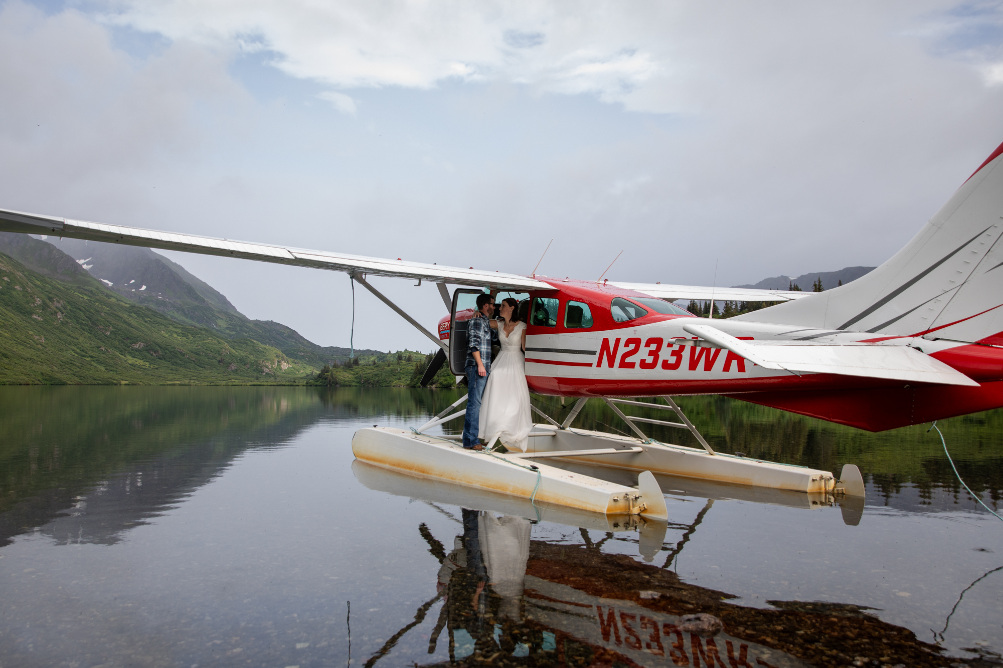 A bride and groom stand on the side of a floatplane that is on a lake in Alaska on their floatplane elopement day.