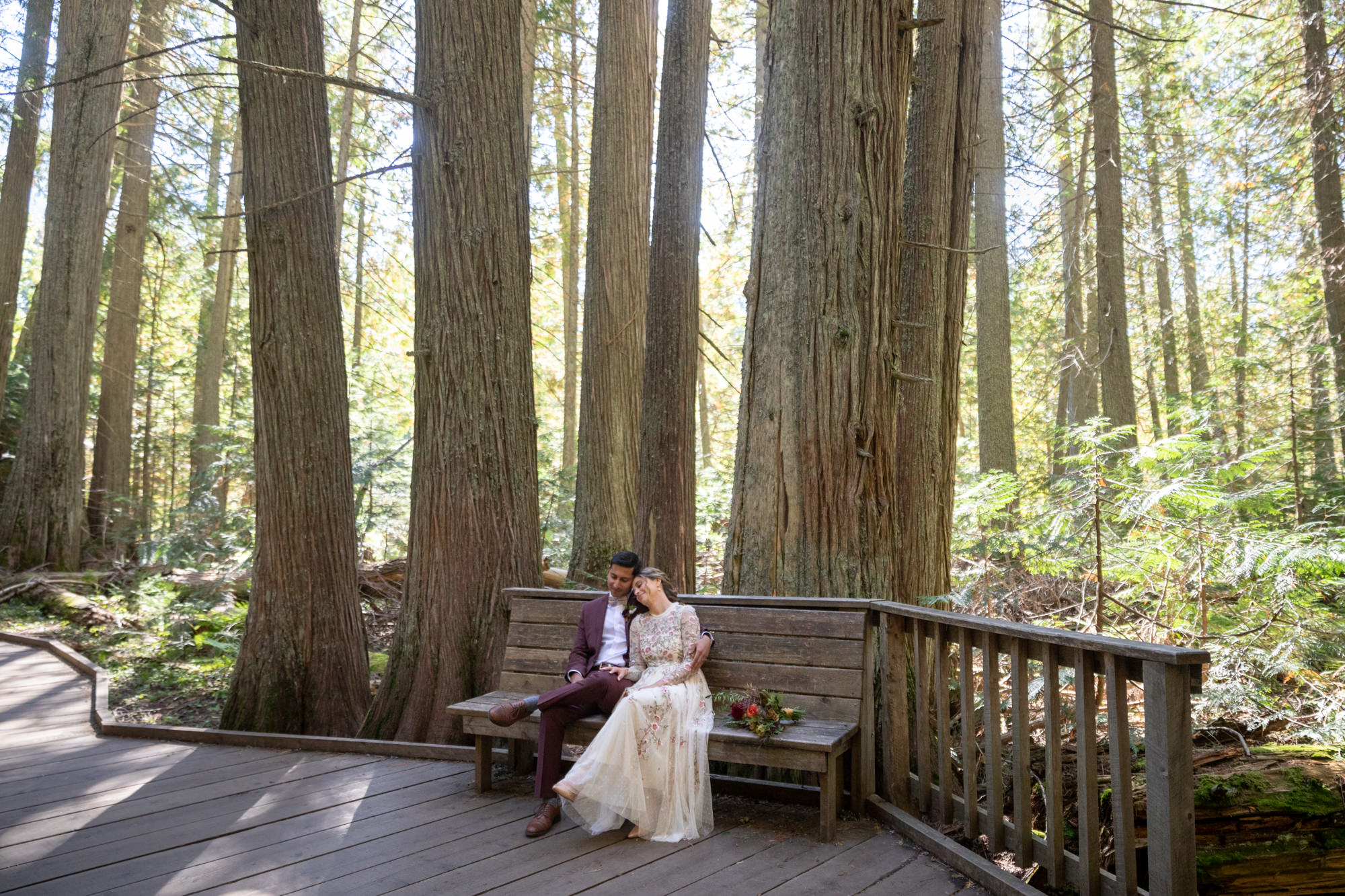 A bride in an offwhite dress with flowers on it, sits on a bench with her groom who is wearing a maroon suit surrounded by tall trees on their forest weddinng day.