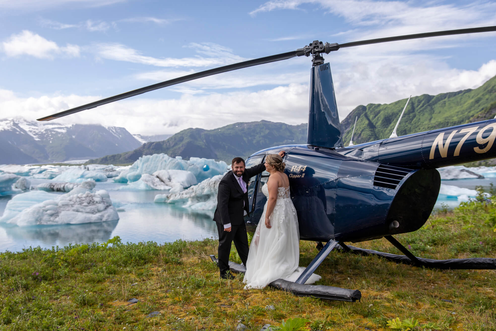 A newlywed couple leans on a helicopter after landing next to a glacier bay full of iceburgs.