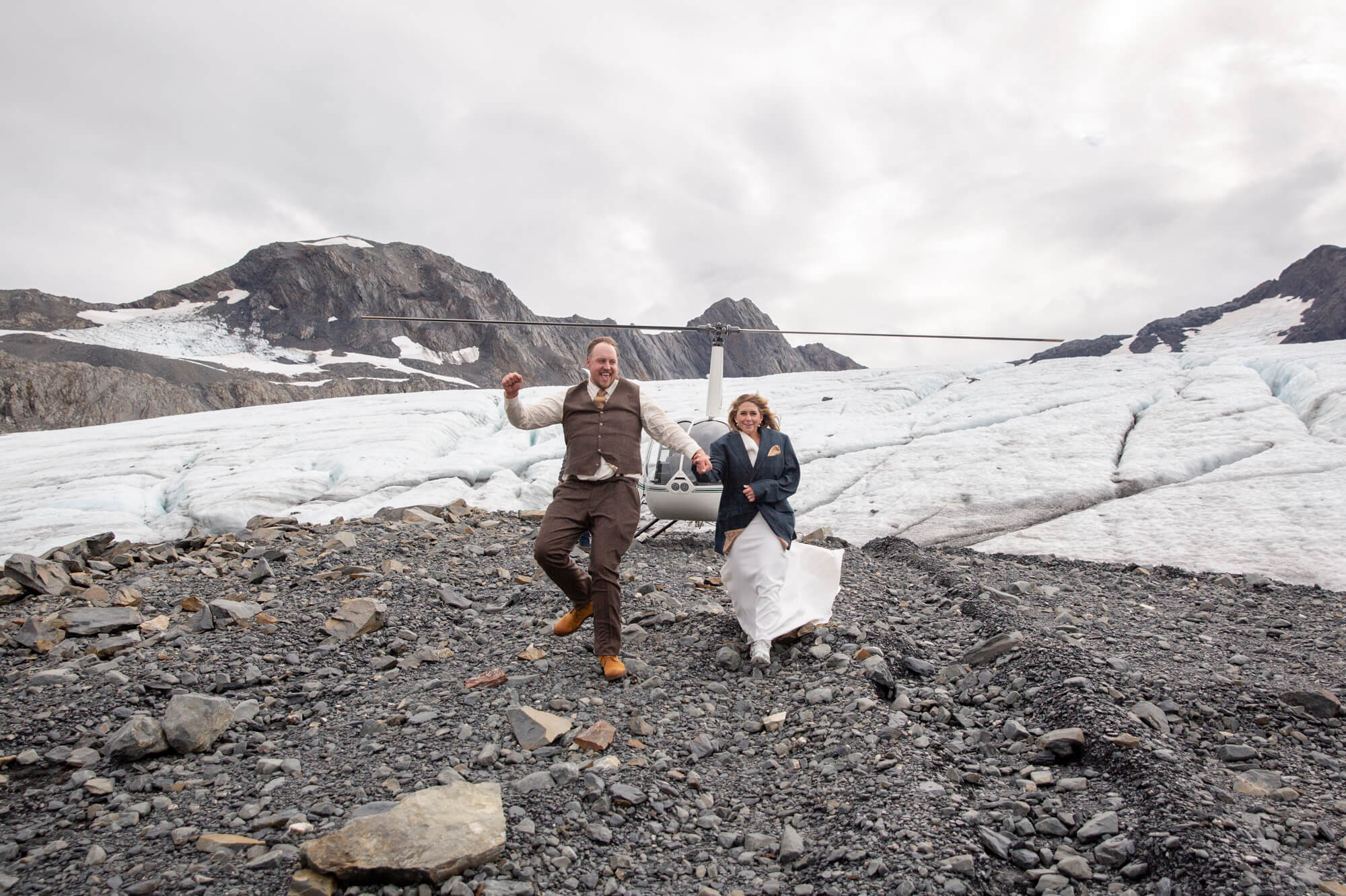 Couple running across rocky terrain in front of a glacier on the Kenai Peninsula, Alaska during an adventurous elopement.
