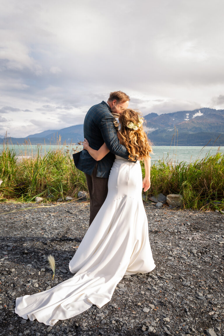 Bride and groom stand next to each other hugging on the beach in Seward Alaska