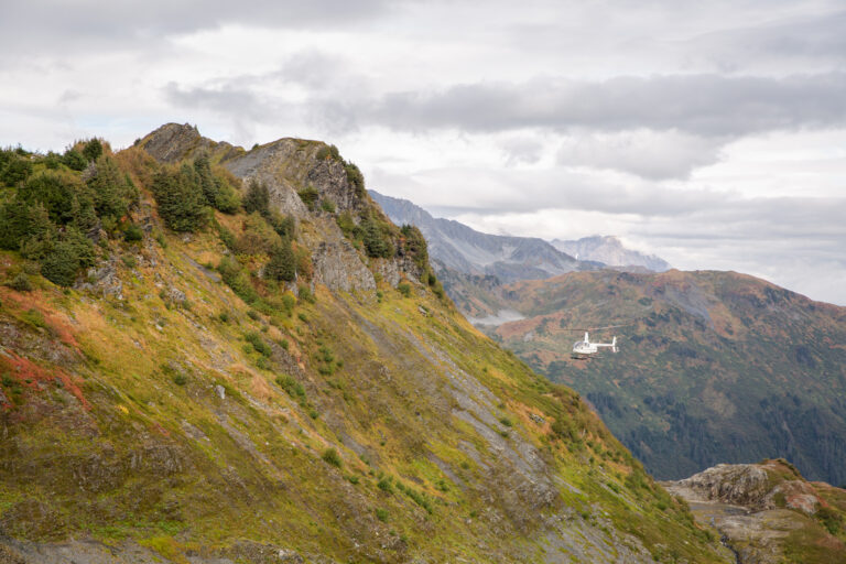 A helicopter in the distance flies over vibrant green mountains in Seward Alaska