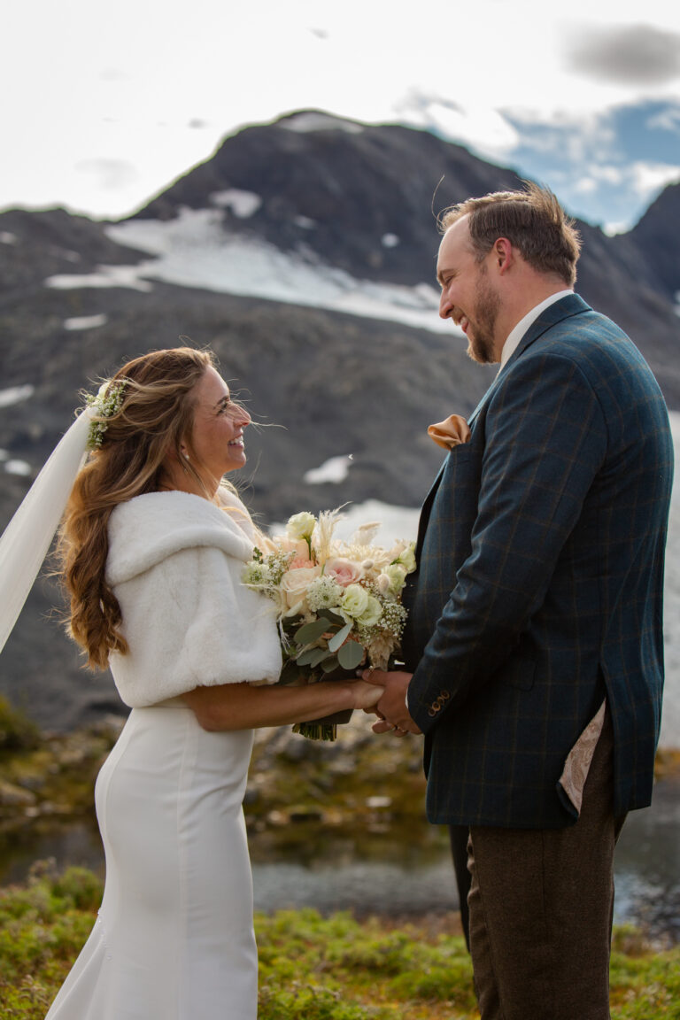 A bride and groom stand facing each other, holding hands and laughing during their elopement ceremony in Seward Alaska.