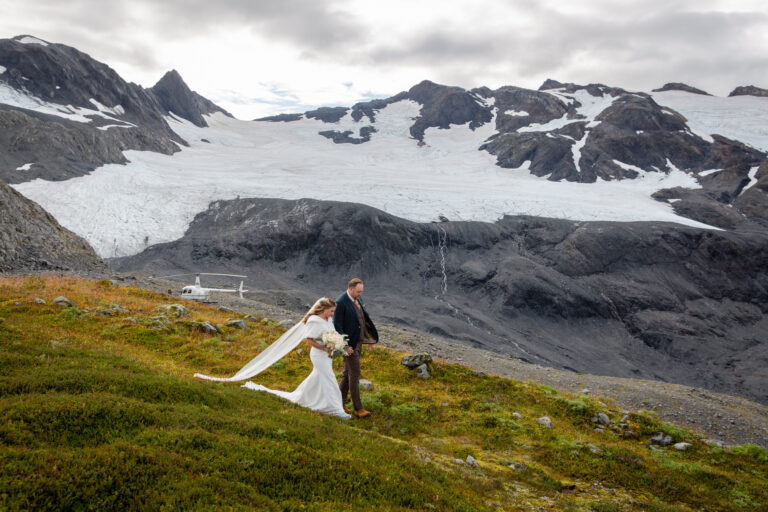 A bride and groom walk hand in hand down a green grassy hill with a helicopter landed on some rocks behind them.