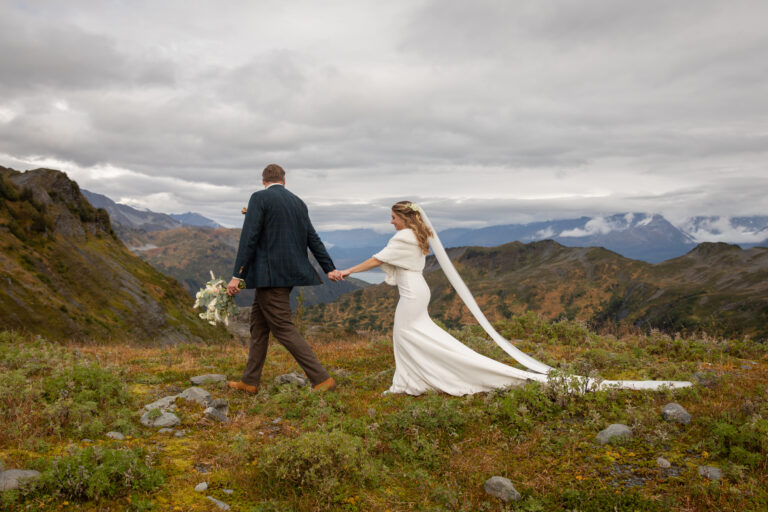 A groom holding a bouquet of flowers leads his bride by the hand across a grassy hill in Alaska.