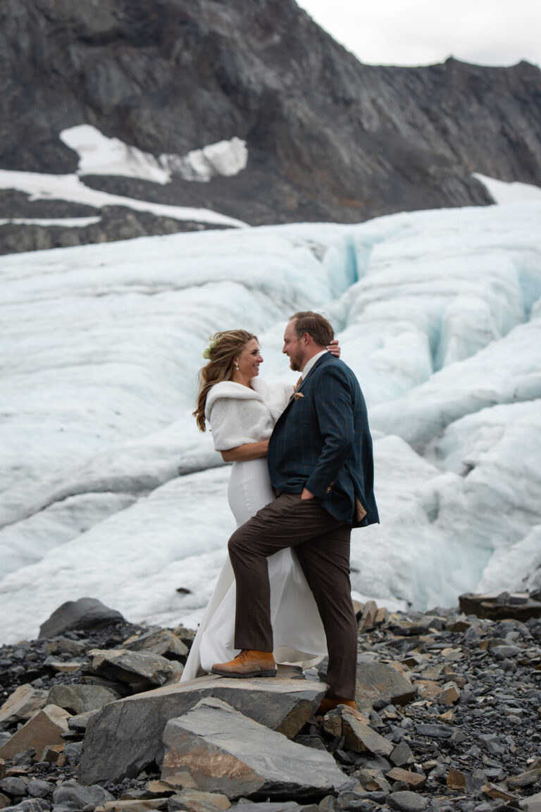 Bride and groom stand on a rock facing each other in an embrace with a glacier behind them.
