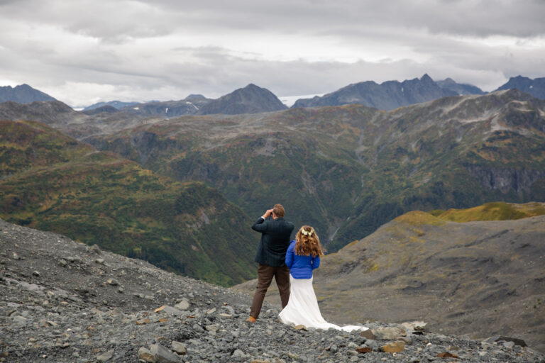Groom faces away from the camera, looking through binoculars while his wife stands next to him wearing a blue jacket over her wedding dress.