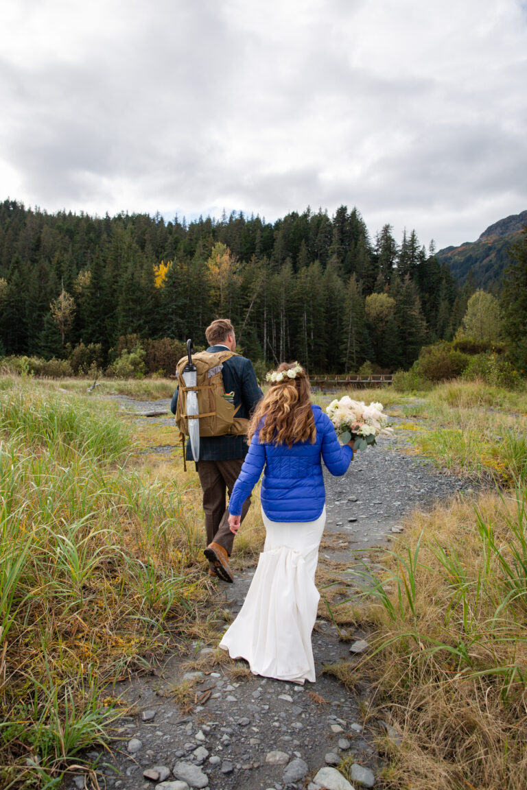 Bride and groom walk along a trail heading towards a forest in front of them.