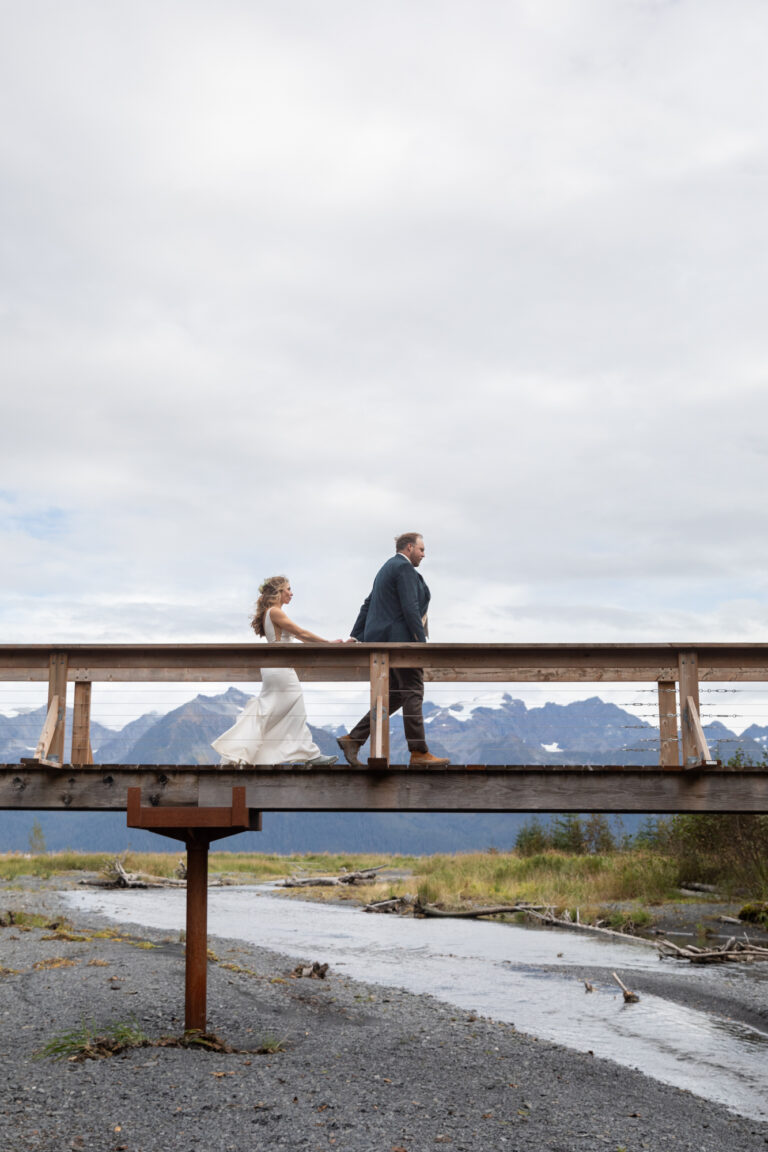 Groom walks in front of bride across a bridge with mountains behind them.