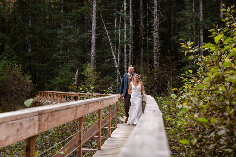Bride and groom walk towards the camera across a bridge with trees behind them.