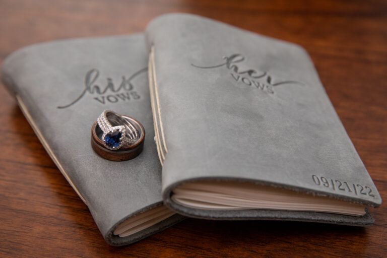 A wedding band and wedding ring with a blue stone rest on top of two gray leather bound vow books.
