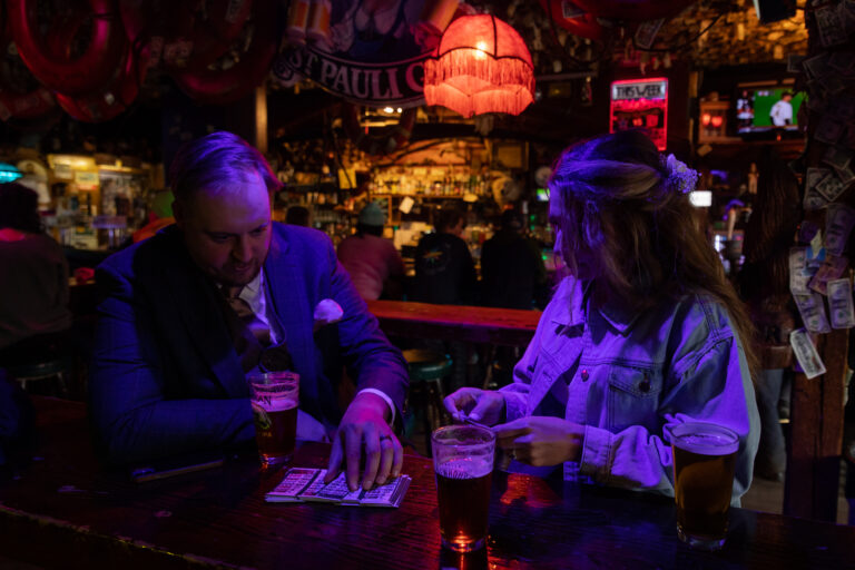 Groom and bride sit in a dive bar with purple, blue lights surrounding them, drinking a cocktail on their wedding day.