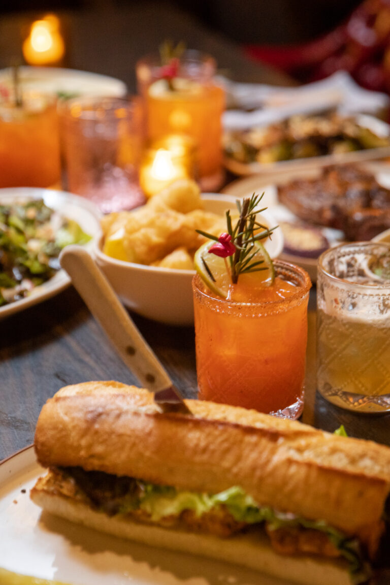 A table full of food with a sandwich on a plate with a knife sticking out of it and an orange cocktail with a sprig of rosemary.