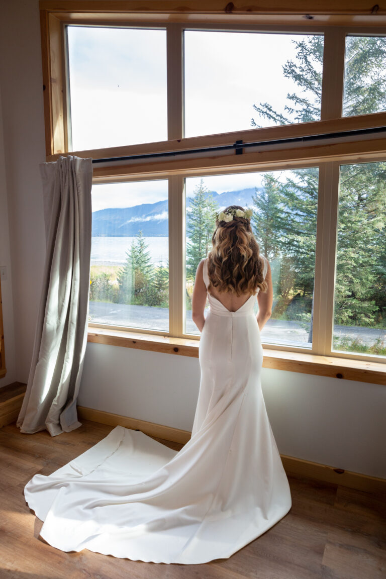 Bride stands looking out the window of her cabin on the morning of her helicopter elopement.