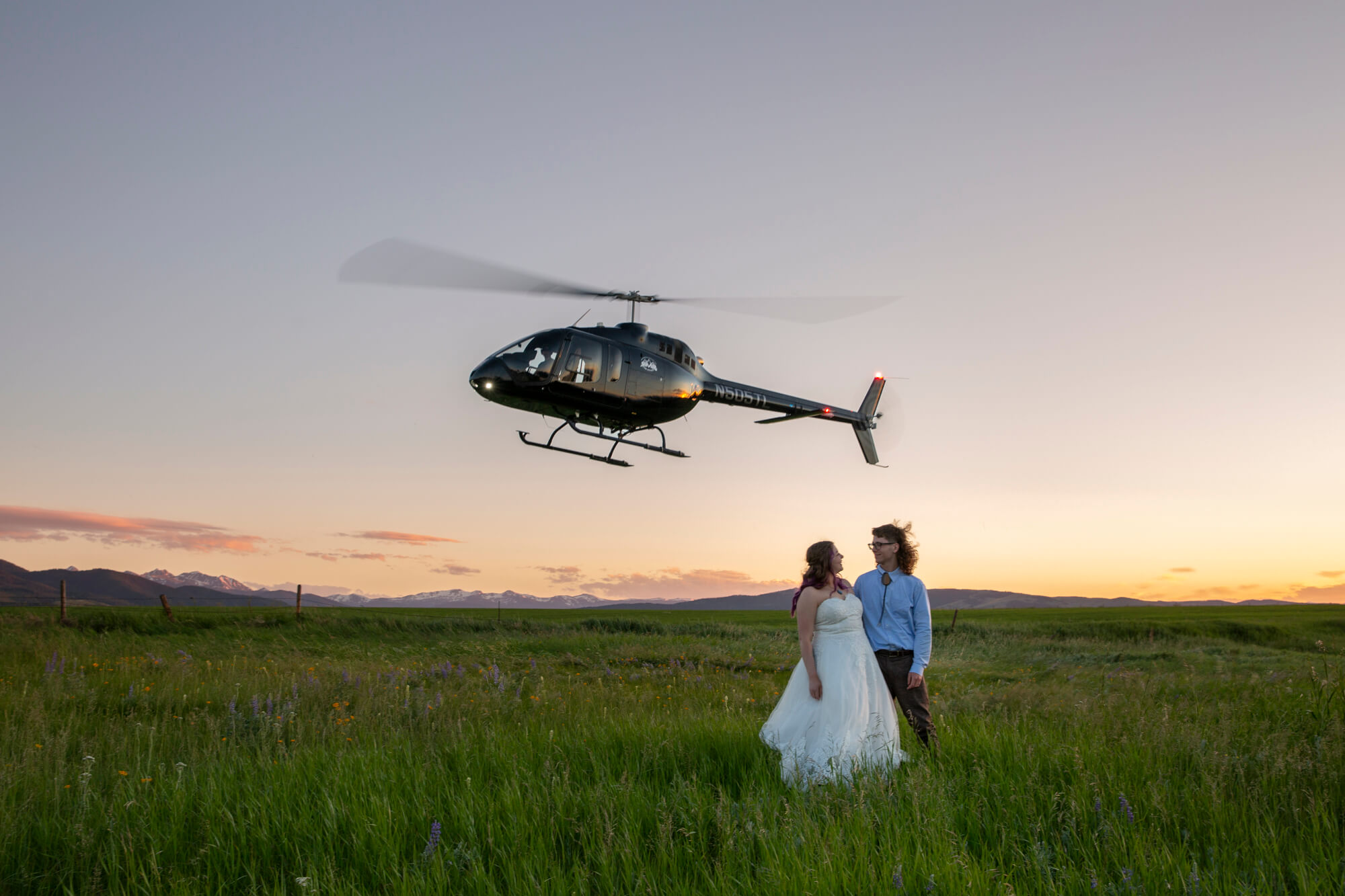7 Best Locations for an Epic Helicopter Elopement in 2025 | Scenic Vows Couple during their Montana helicopter elopement at sunset as the helicopter lifts off in a mountain meadow.