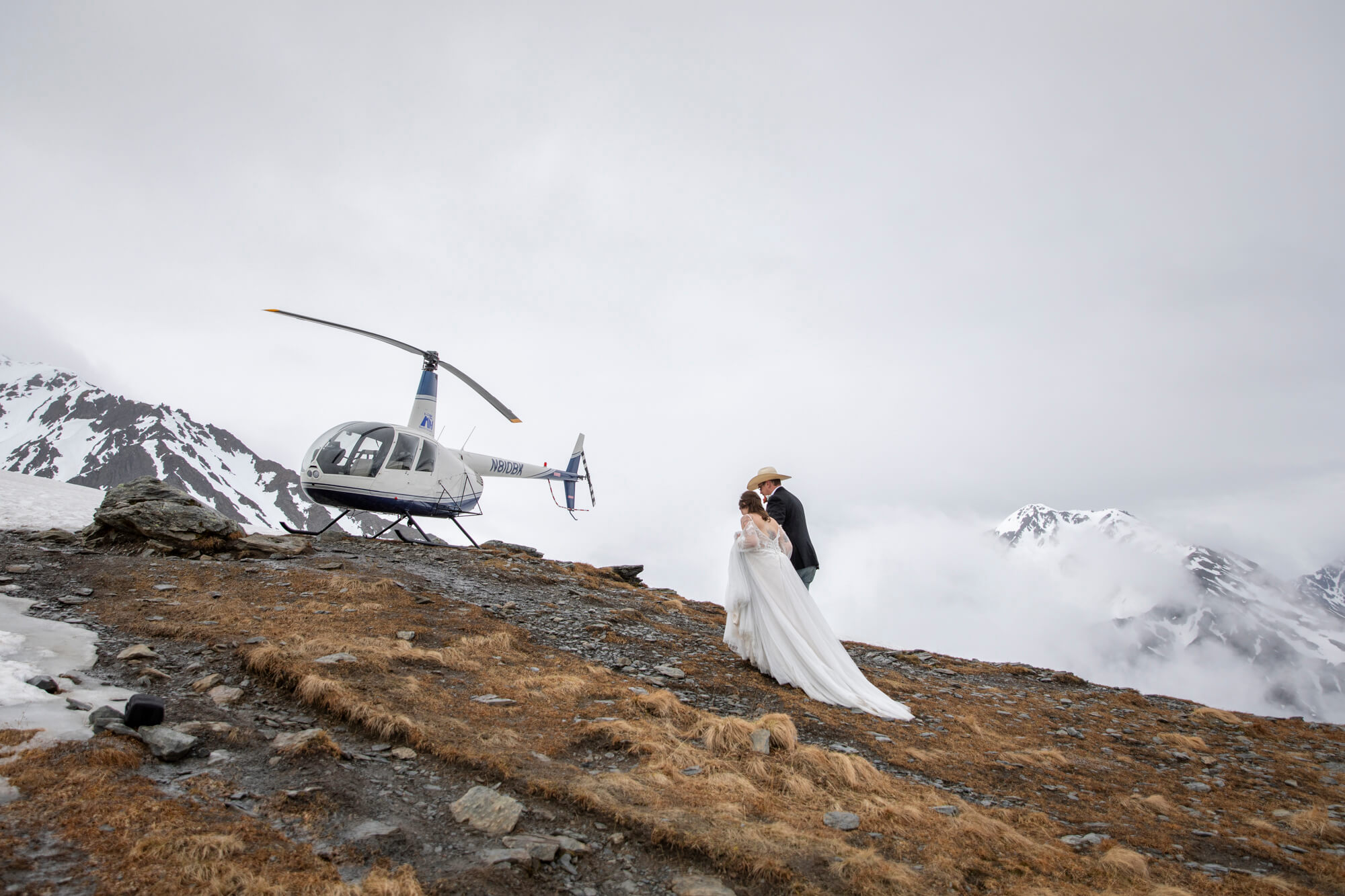 Bride and groom walk along a rocky ridgeline near a white helicopter, surrounded by clouds and snowy peaks after their helicopter elopement.