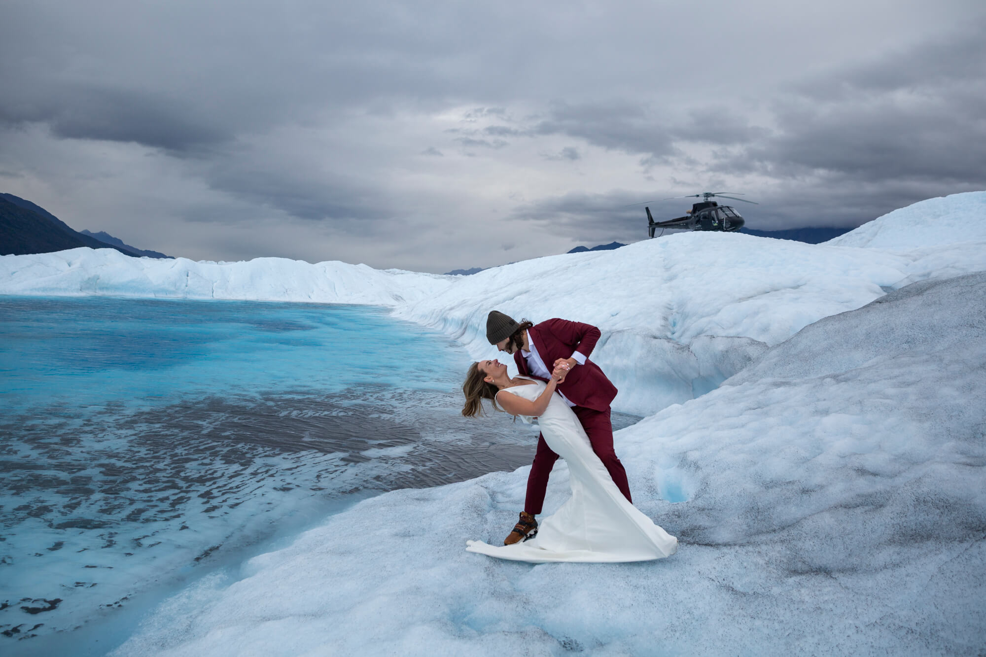A man dips his new bride next to a bright blue glacier pool, their helicopter is parked on the glacier behind them. 