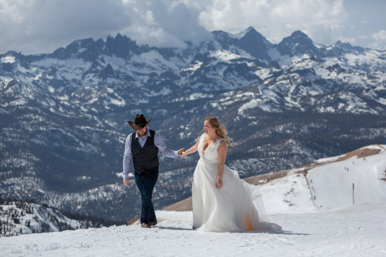 A bride and groom walk up a snowy hill hand in hand with giant mountains behind them.